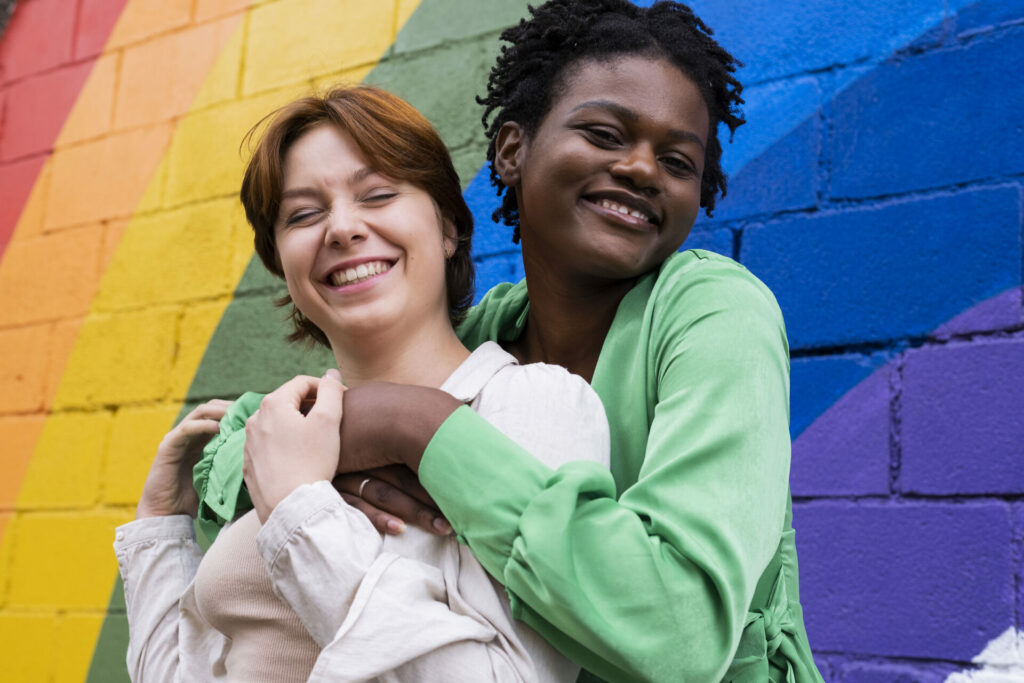 Glückliche Frau, die ihre Freundin vor einer an die Wand gemalten Regenbogenflagge umarmt